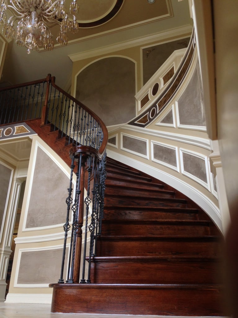 Elegant curved wooden staircase with ornate black iron railing in a classical hallway with cream and beige walls, decorated ceiling, and crystal chandelier