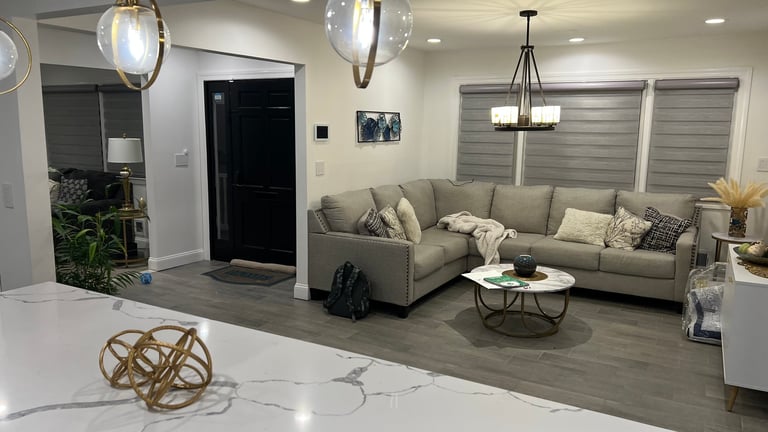 Modern living room with beige sectional sofa, marble kitchen island in foreground, pendant lighting, and gray wood flooring